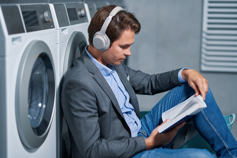 A man is reading a physical book while listening to an audiobook in the laundry room